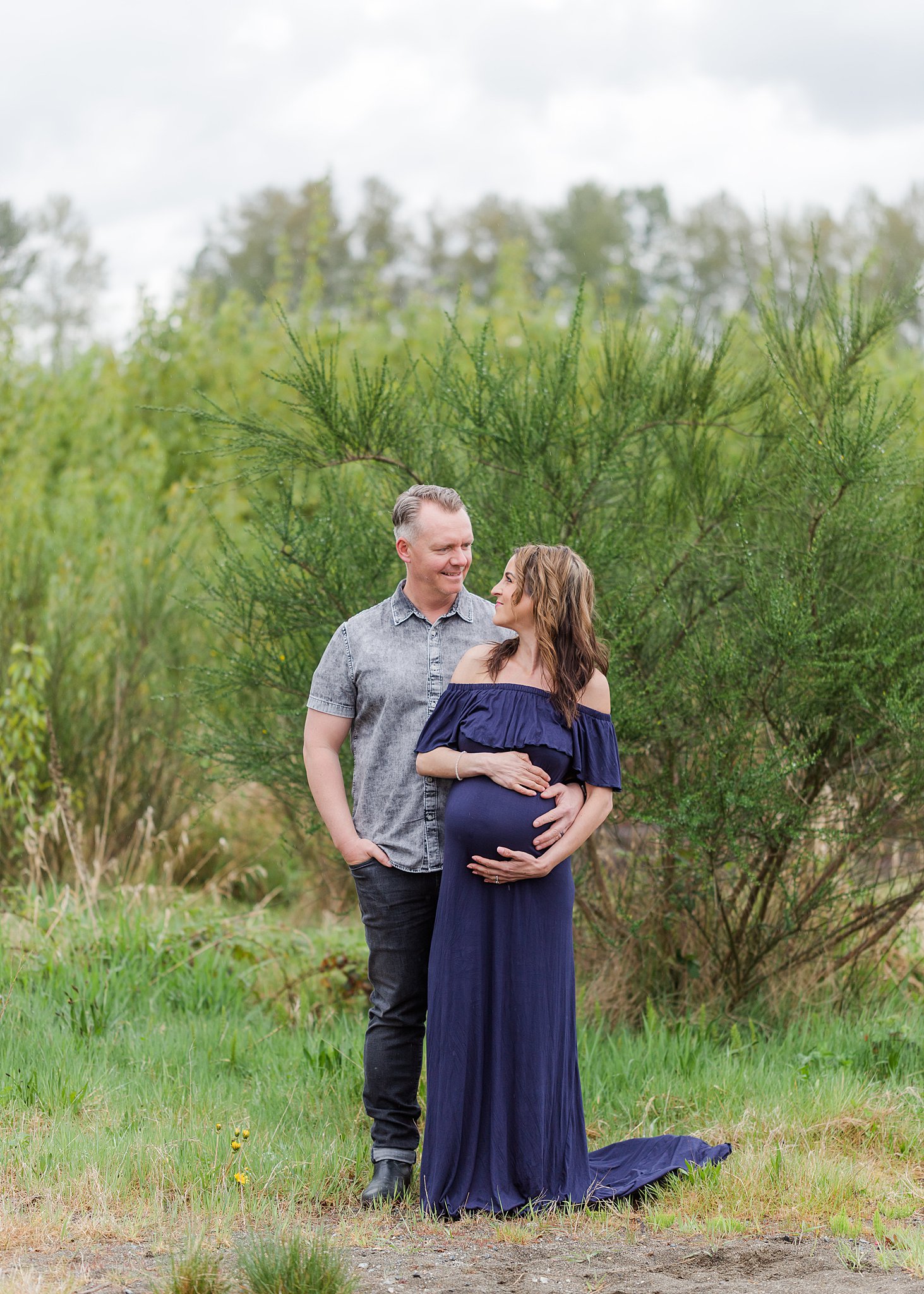 A mother to be in a blue maternity gown is hugged by her husband beside a park path