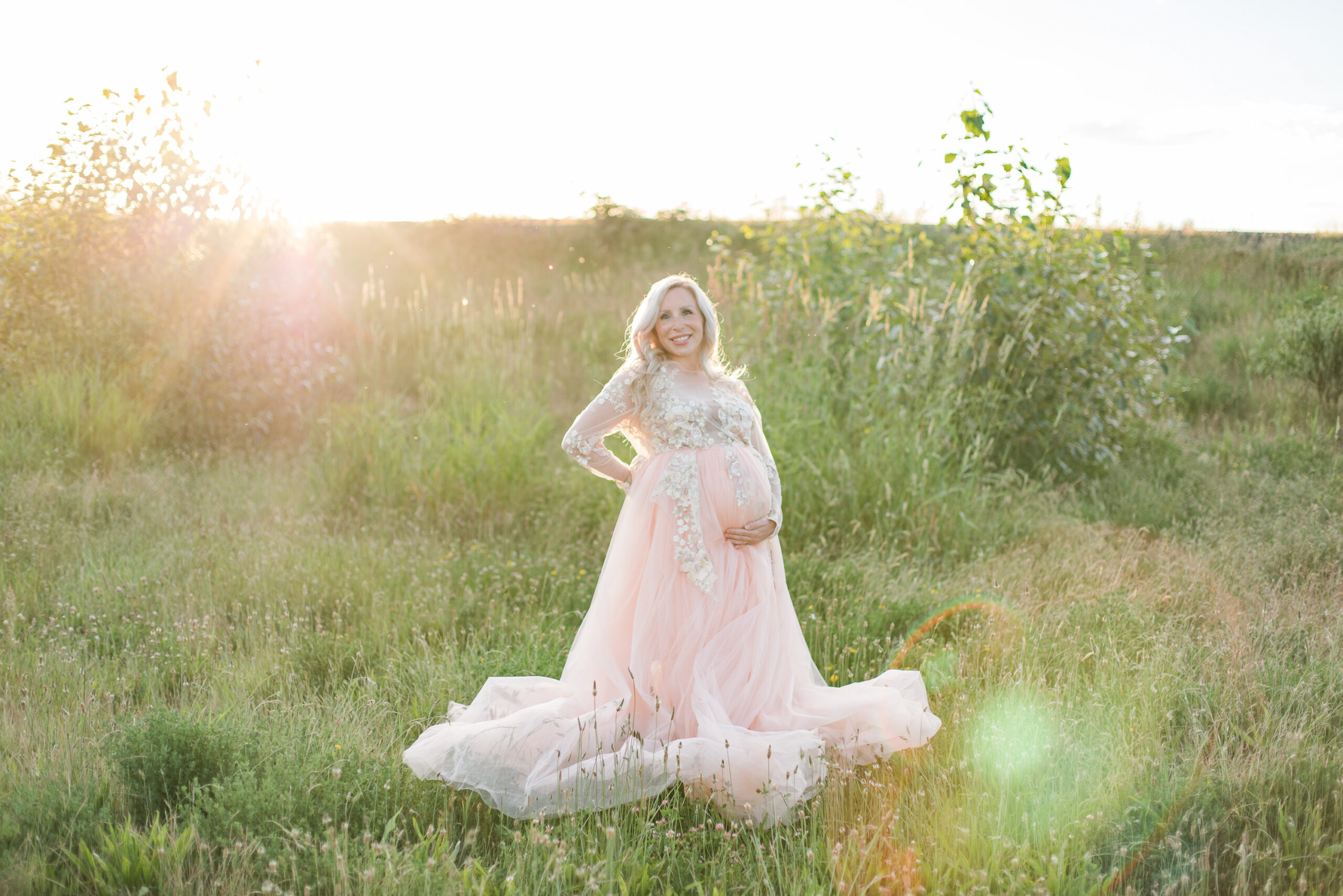 A pregnant woman stands in a grassy green field with a hand on her bump in a flowing pink maternity gown